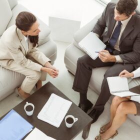 Realtor sitting on a couch discussing home-selling plans with a husband and wife during an interview.
