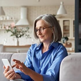 Older woman sitting on a couch using her mobile phone, considering how to contact a realtor.